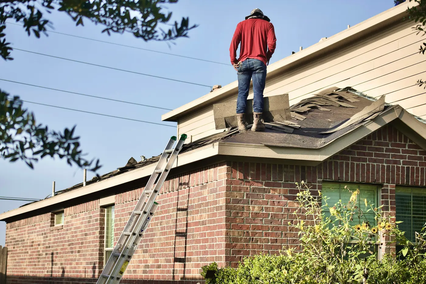 A person standing on a house with a damaged roof, a ladder leaning against it